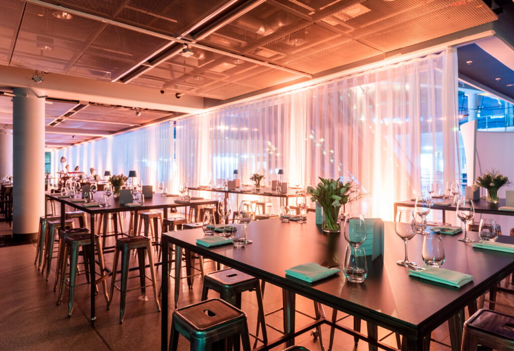 Modernes Restaurant mit hohen Tischen, Hockern, Gläsern und sanfter Beleuchtung hinter transparenten Vorhängen.