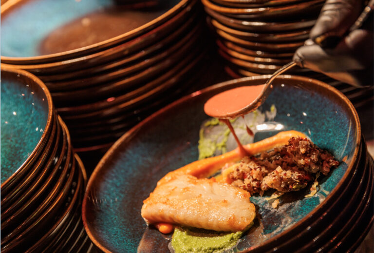 Eine behandschuhte Hand beträufelt ein mit Blattgemüse und knackigem Gemüse angerichtetes Fischgericht mit Sauce, frisch zubereitet im Station Airport für Veranstaltungen.