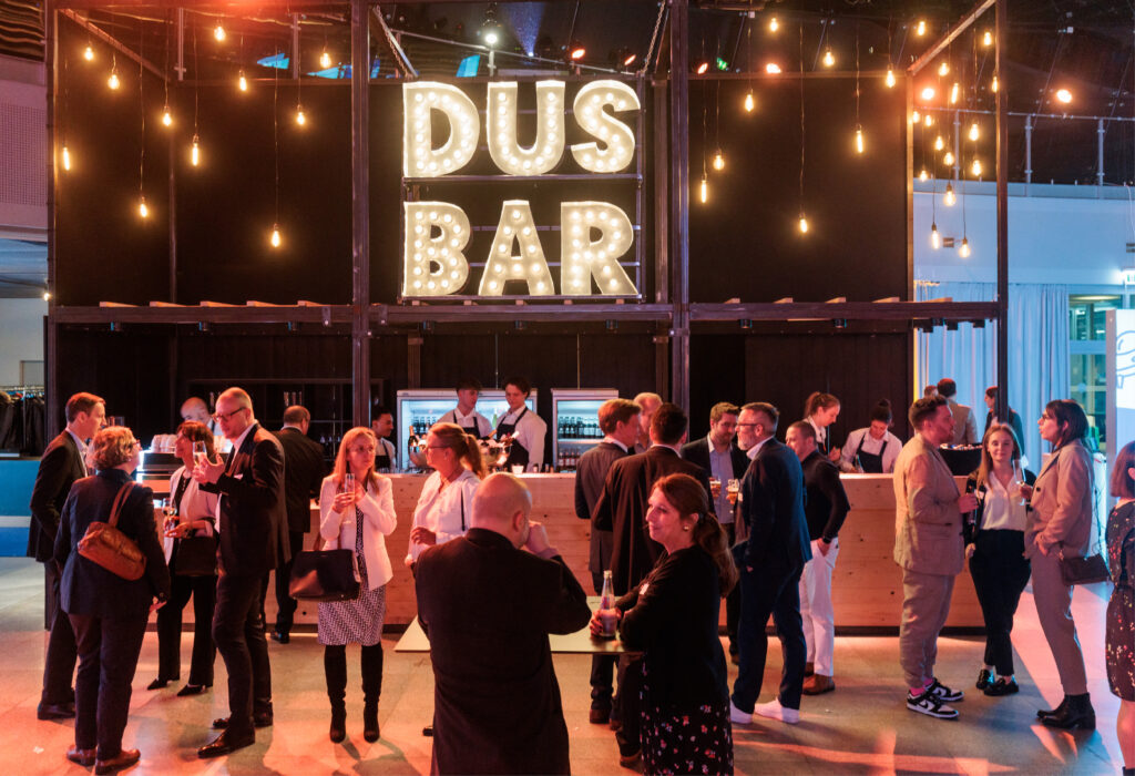 Bei einem Bar-Event unter einem großen beleuchteten DUS BAR-Schild knüpfen die Leute Kontakte und tauschen sich aus.