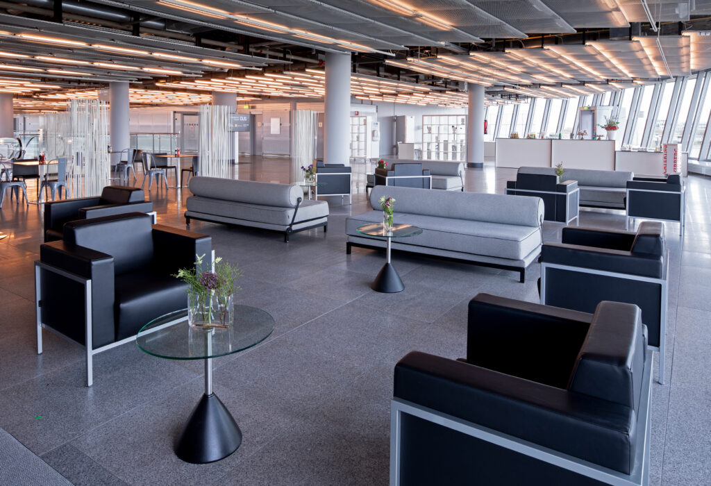 Moderner Loungebereich in der Station Airport für Veranstaltungen mit schwarzen Stühlen, grauen Sofas, Glastischen und großen Fenstern in einem geräumigen, hellen Raum.