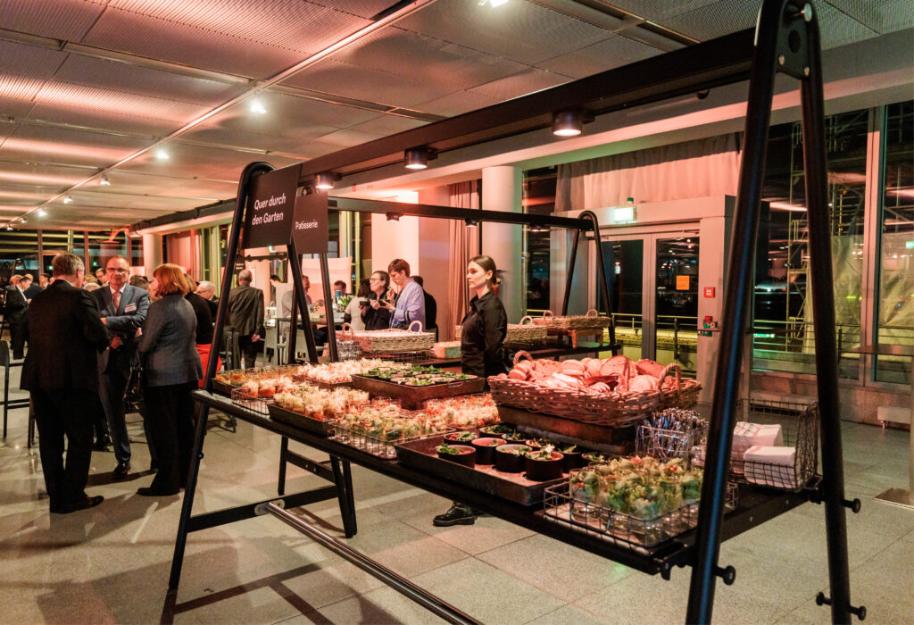Ein Buffettisch mit verschiedenen Speisen bei einer Indoor-Veranstaltung, wie sie beispielsweise in der Station Airport für Veranstaltungen stattfindet, bei der im Hintergrund geselliges Beisammensein herrscht.