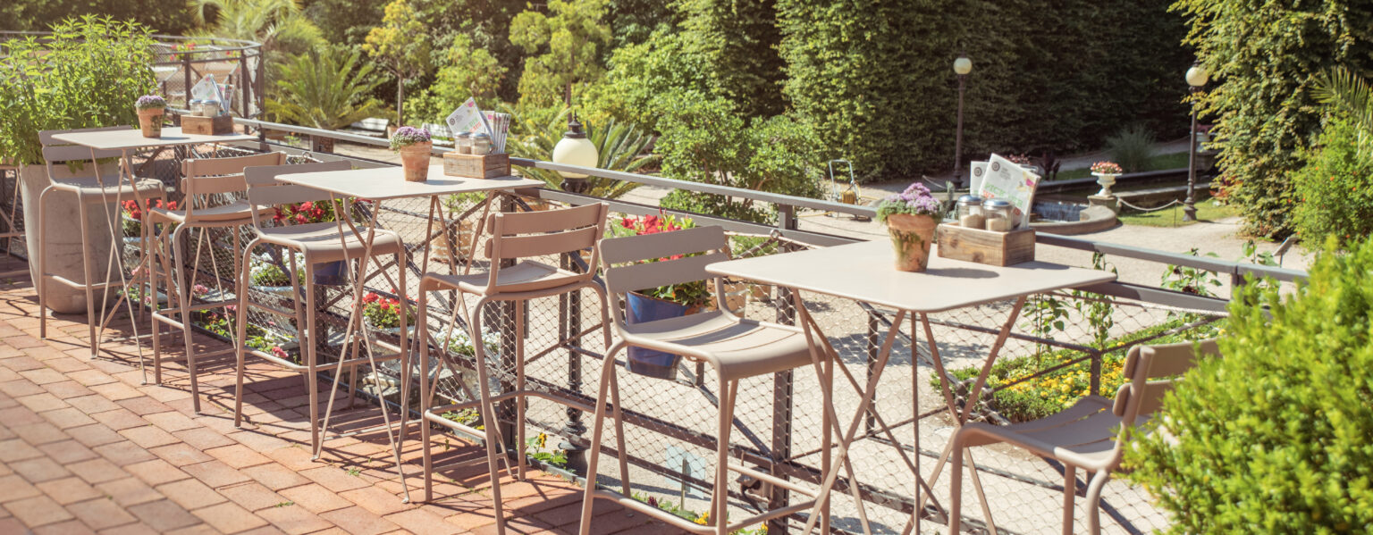 Café-Terrasse im Freien mit leeren Tischen und Stühlen, Topfpflanzen und Blick auf den Garten im Sonnenlicht – ideal zum Entspannen oder Genießen des Caterings im Dank Augusta.