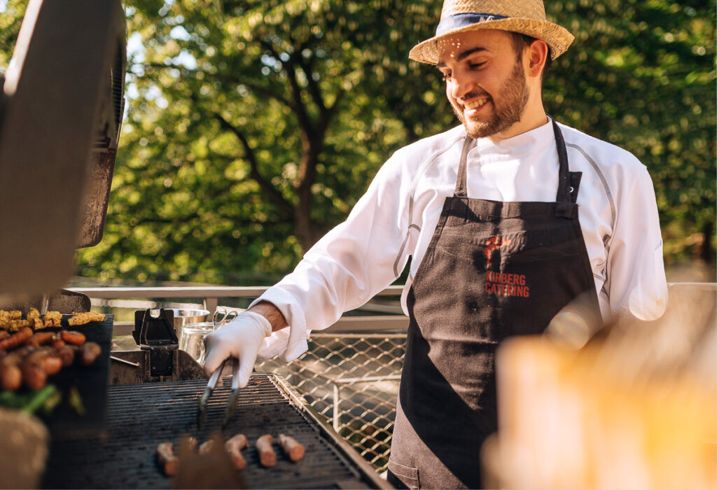 Ein lächelnder Koch mit Strohhut grillt an einem sonnigen Tag im Freien Würstchen und Gemüse und zeigt die Freude am Catering im Dank Augusta.