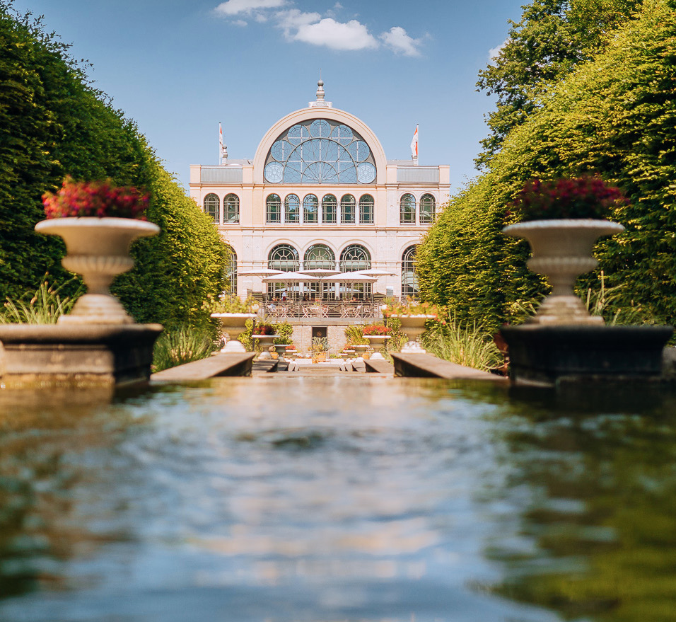 Hinter gepflegten Hecken erhebt sich ein großes Glasgebäude mit einem spiegelnden Brunnen und Pflanzgefäßen im Vordergrund – eine ideale Kulisse für Veranstaltungen mit Catering im Dank Augusta.