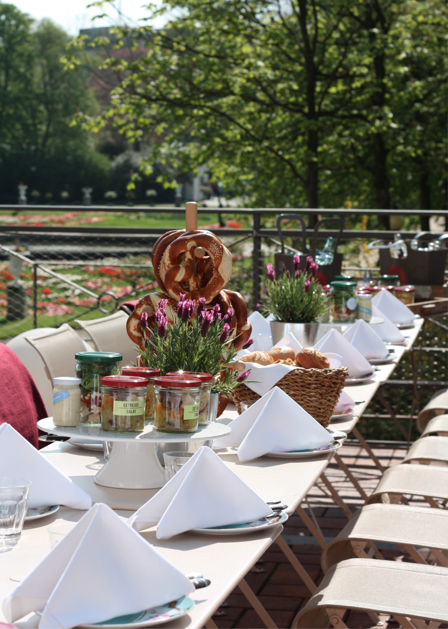 Tisch im Freien mit weißen Servietten, Blumen, Gläsern und Brezeln, umgeben von Stühlen und Grünpflanzen – perfekt für Catering-Treffen im Dank Augusta.