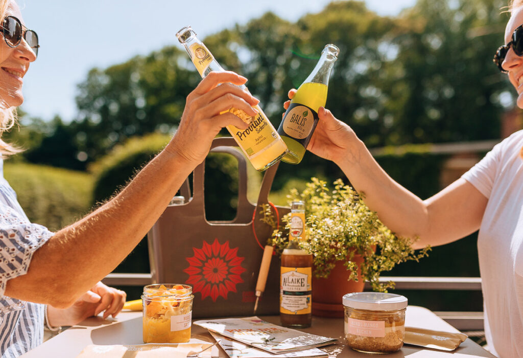 Zwei Menschen stoßen im Freien mit Flaschengetränken an und genießen Snacks und Sonnenlicht auf einem Tisch.