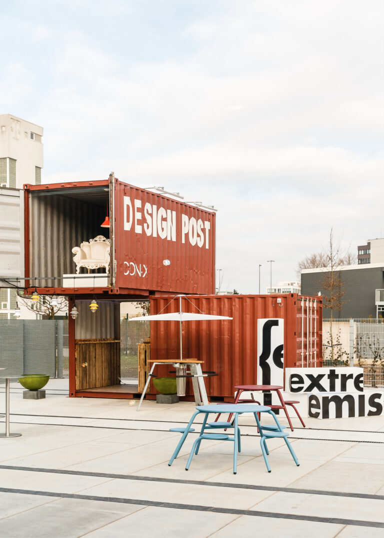 Rote Schiffscontainer, gestapelt als moderner Ausstellungsraum im Freien mit der Aufschrift DESIGN POST und ausgestellten Möbeln, präsentieren Design Post als Eventlocation.