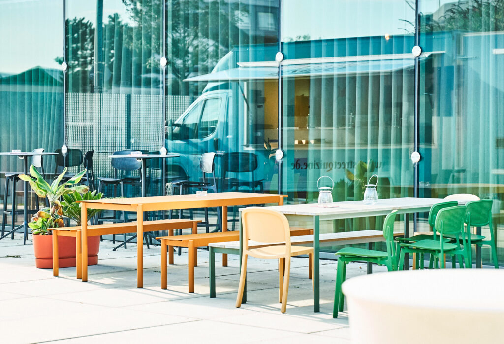 Bunte Tische und Stühle im Freien stehen leer neben einem Glasgebäude und einem geparkten Foodtruck und machen die Design Post als Eventlocation zu einem einladenden Ort für Zusammenkünfte.