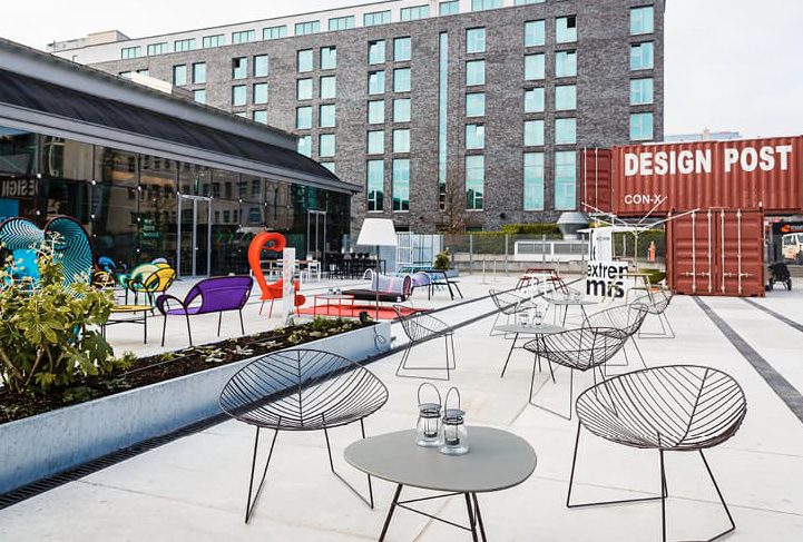 Außenterrasse mit modernen Drahtstühlen und farbenfrohen Möbeln, neben einem Backsteingebäude und einem DESIGN POST Container – perfekt für Treffen in der Design Post als Eventlocation.