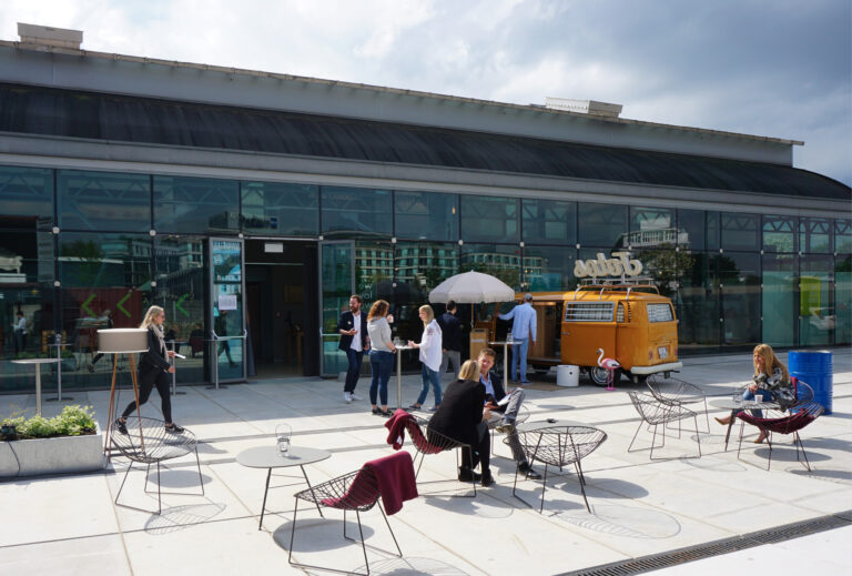 An einem sonnigen Tag sitzen Menschen vor einem modernen Gebäude – Design Post als Eventlocation – mit einem gelben Imbisswagen und Drahtstühlen und unterhalten sich.