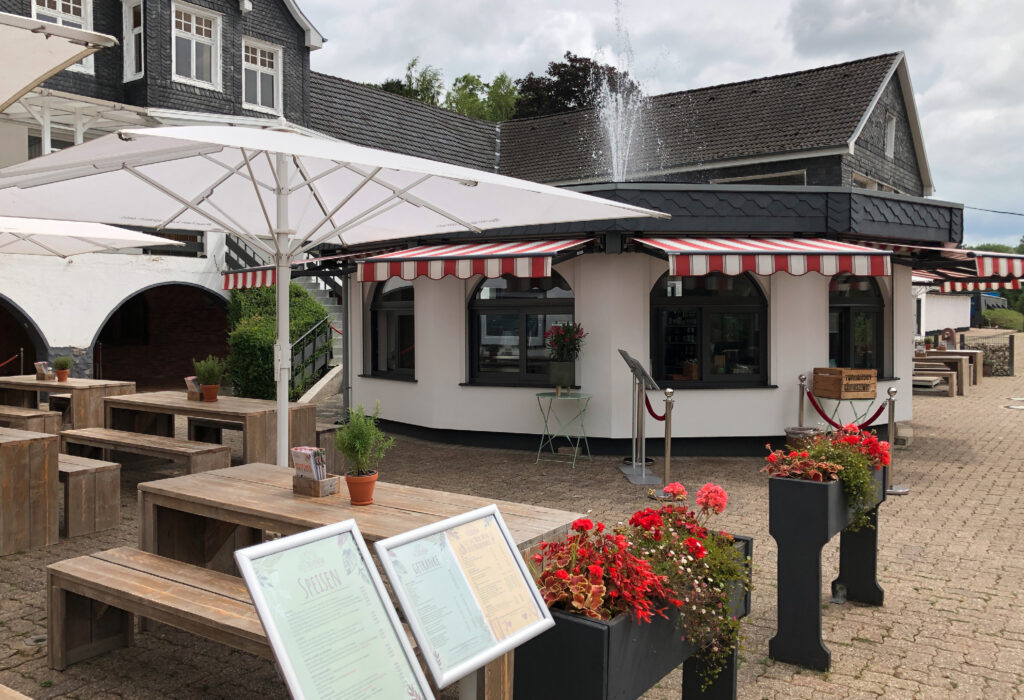 Straßencafé mit Holztischen, Blumenkübeln, Sonnenschirmen und einem kleinen weißen Gebäude mit rot gestreiften Markisen – perfekt für Zusammenkünfte im Haus Diepental für Feiern.