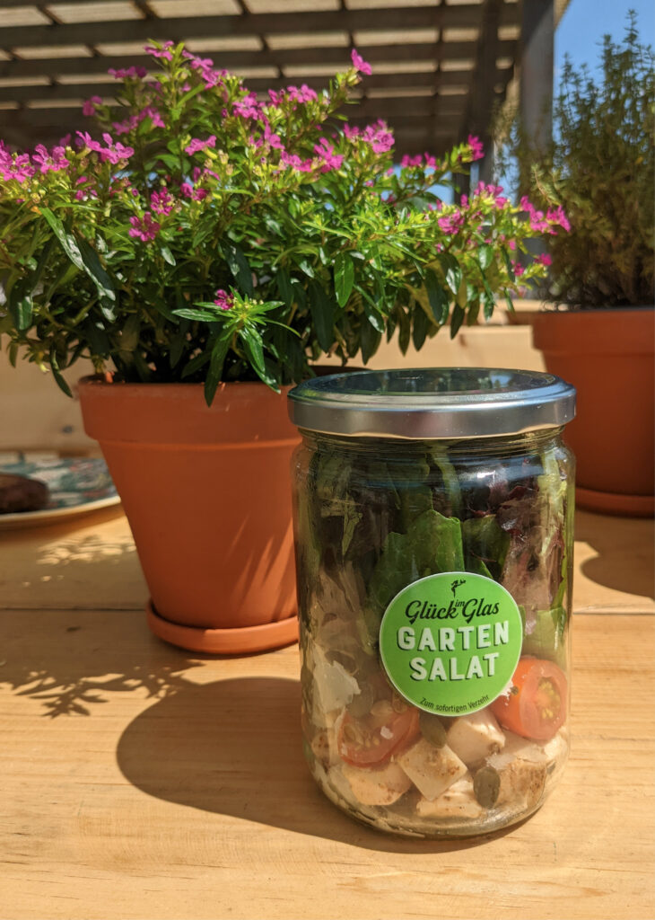Ein Glas mit geschichtetem Gartensalat steht auf einem Holztisch neben blühenden Topfpflanzen im Sonnenlicht – eine einladende Szene, die an Zusammenkünfte im Haus Diepental für Feiern erinnert.
