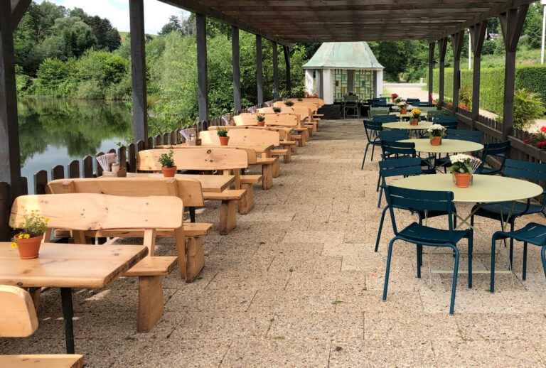 Außensitzbereich mit Holzbänken, runden Tischen, Stühlen und Topfpflanzen an einem See unter einem Baldachin – perfekt für Versammlungen oder Veranstaltungen im Haus Diepental für Feiern.