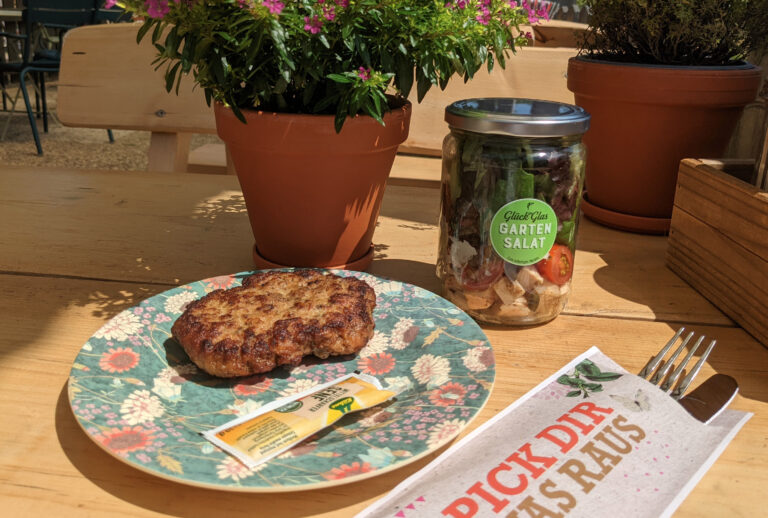 Ein Teller mit Blumenmuster, einem Frikadellenstück und Senf, einem Glas Salat und Besteck auf einem Holztisch im Freien – perfekt für Zusammenkünfte im Haus Diepental für Feiern.