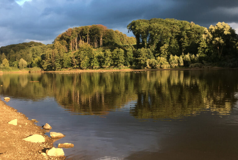 Bäume spiegeln sich in einem ruhigen See unter einem teilweise bewölkten Himmel, im Vordergrund säumen Felsen das Ufer – eine perfekte Landschaft in der Nähe des Haus Diepental für Feiern.