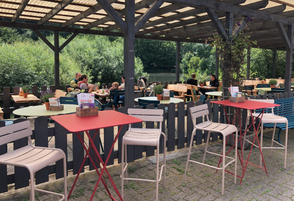 Straßencafé mit roten Tischen, leeren Stühlen und Menschen, die an Tischen unter einer Holzpergola sitzen – ein einladender Ort im Haus Diepental für Feiern.