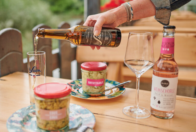 Eine Hand gießt Bier in ein Glas neben Salatgläsern und einer Weinschorle auf einem Holztisch im Haus Diepental für Feiern.
