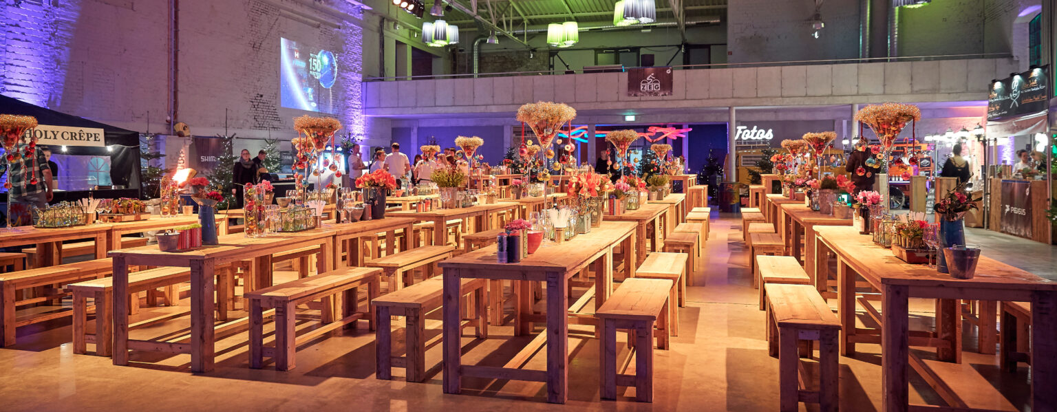 In einem großzügigen, dekorierten Veranstaltungssaal im Dock 2 in Köln für Events sind mit bunten Blumen geschmückte Holztische und Bänke aufgestellt, und die Leute mischen sich untereinander.