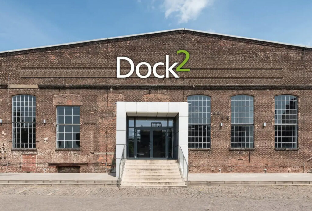 Backsteingebäude mit großen Fenstern und einem Schild mit der Aufschrift Dock2 über der Eingangstreppe unter blauem Himmel – das Dock 2 in Köln für Events ist der perfekte Veranstaltungsort für unvergessliche Anlässe.