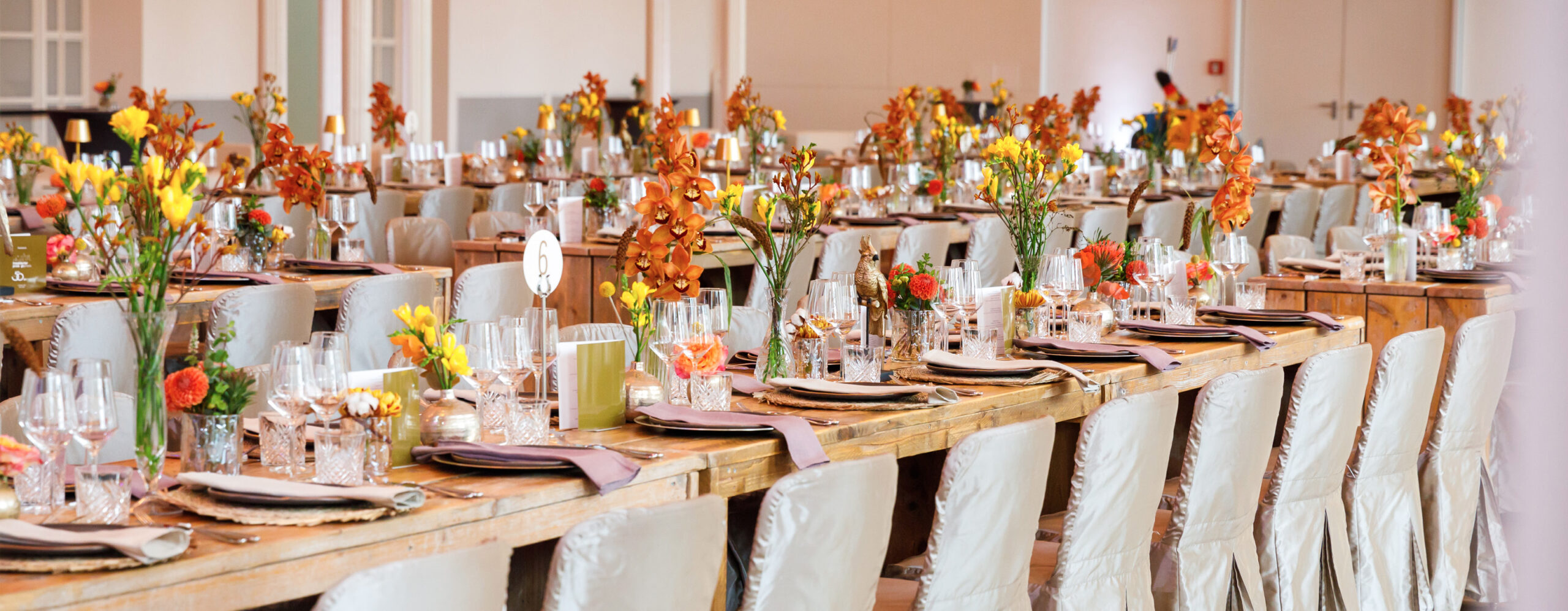 Lange Banketttische für eine Veranstaltung, dekoriert mit orange-gelben Blumen und elegantem Geschirr - perfekt arrangiert für Catering in der Flora Köln.
