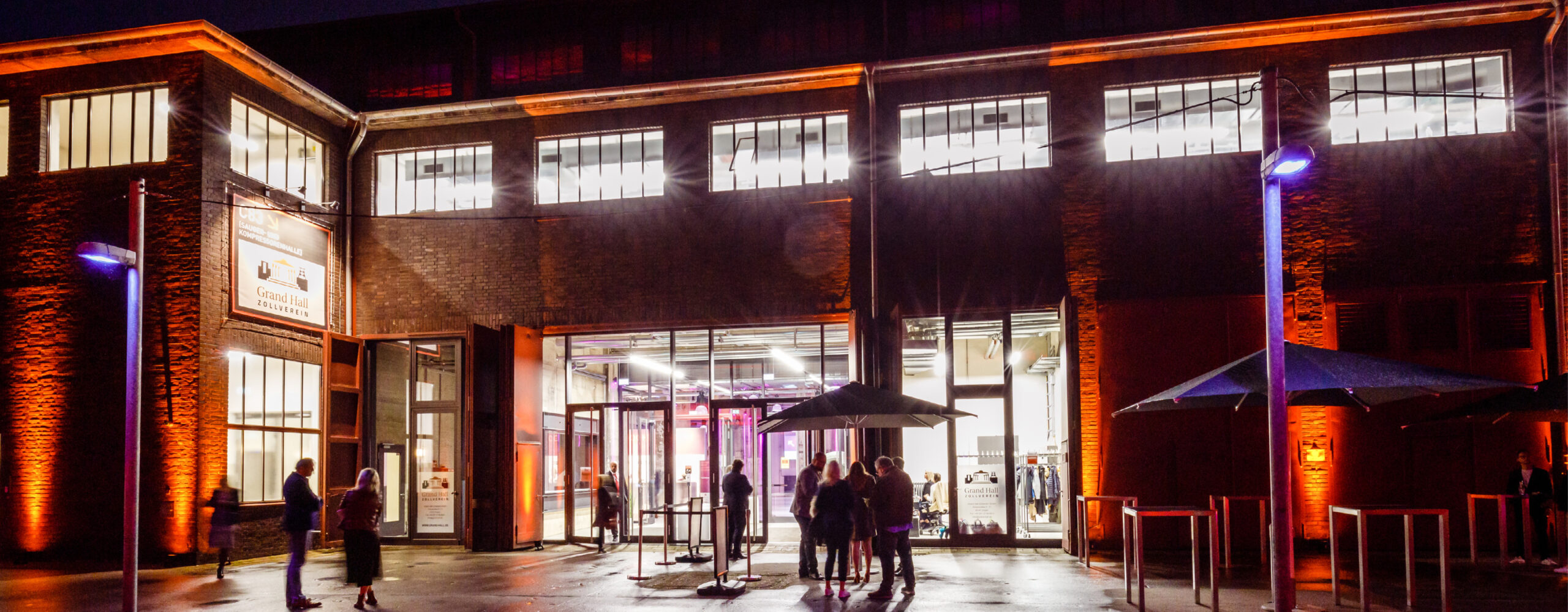 Vor einem hell erleuchteten, modernen Gebäude mit Glastüren versammeln sich nachts Menschen unter orangefarbenem und violettem Licht – vielleicht genießen sie während einer eleganten Abendveranstaltung das Catering in der Grand Hall Zollverein.