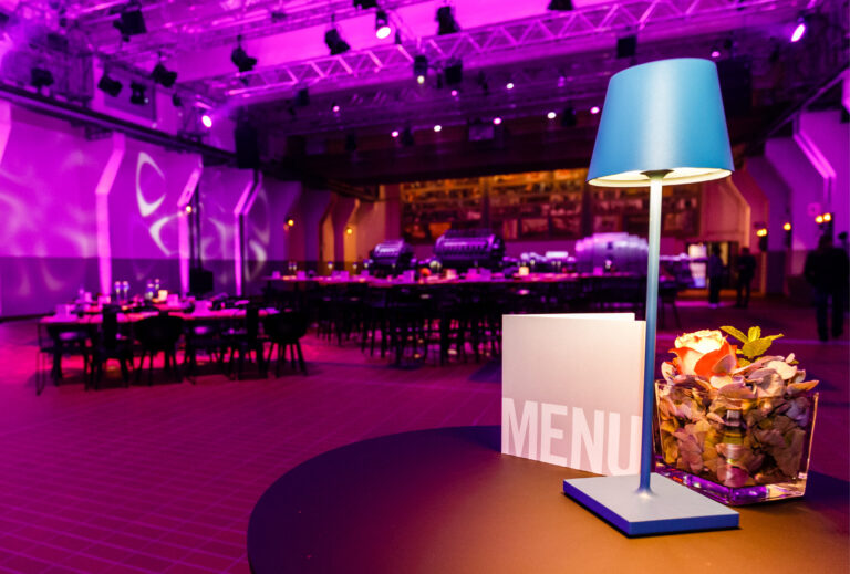Tisch mit blauer Lampe, Menü und Tafelaufsatz in einem violett beleuchteten Veranstaltungsraum mit Tischen im Hintergrund, perfekt für das Catering in der Grand Hall Zollverein.