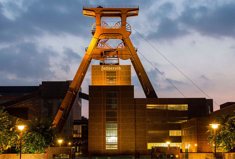 Eine große beleuchtete Industriehalle mit Zollverein-Schriftzug vor einem bewölkten Abendhimmel – die perfekte Kulisse für das Catering in der Grand Hall Zollverein.
