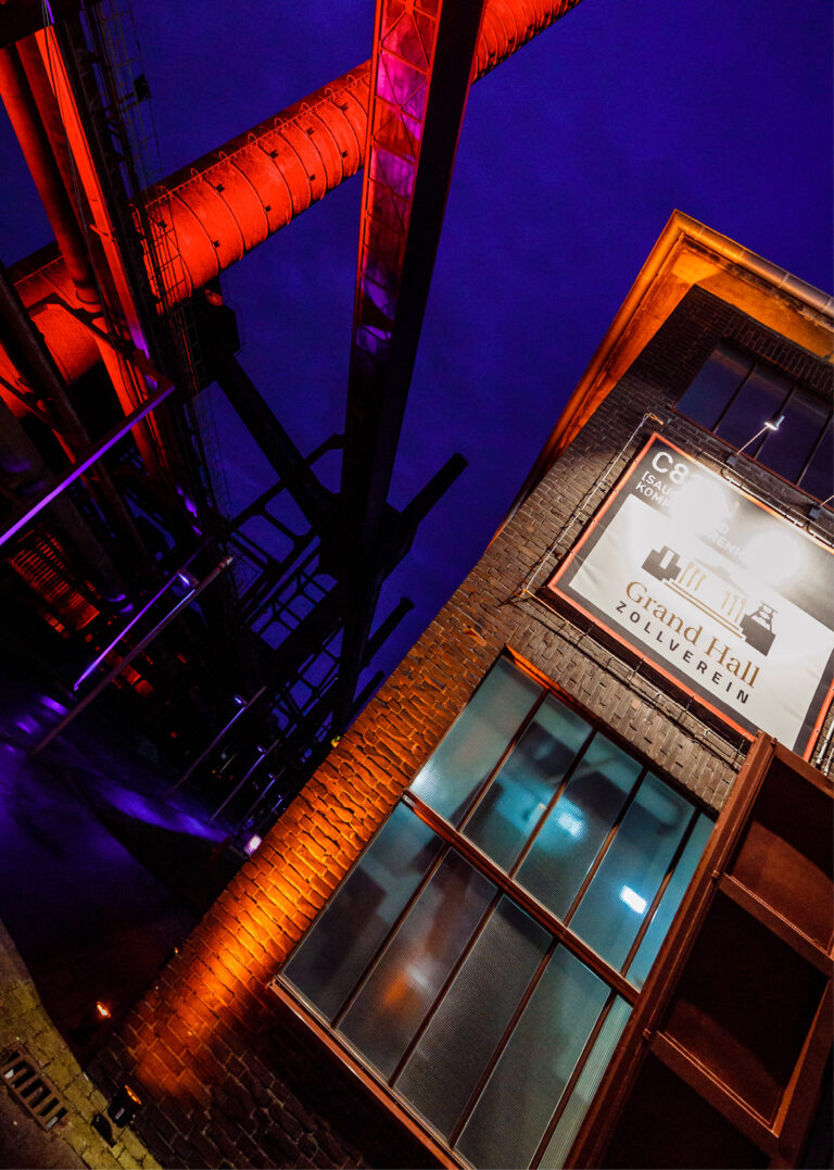 Backsteingebäude mit großen Fenstern und Industrierohren, die nachts von orangefarbenem Licht beleuchtet werden. Auf dem Schild steht „Grand Hall Zollverein“, ein beeindruckender Veranstaltungsort, der für exklusive Events und Catering in der Grand Hall Zollverein bekannt ist.