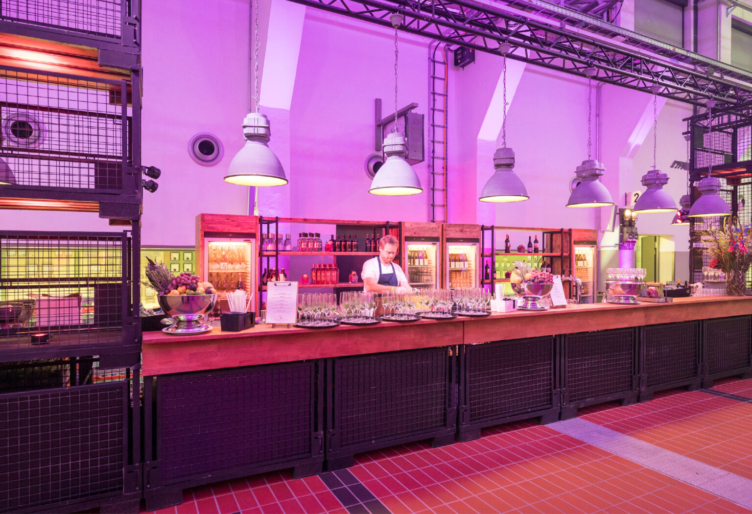 Eine hell erleuchtete Bar mit einem Barkeeper, der hinter einer Holztheke unter Hängelampen Drinks zubereitet und so das stilvolle Ambiente des Caterings in der Grand Hall Zollverein einfängt.