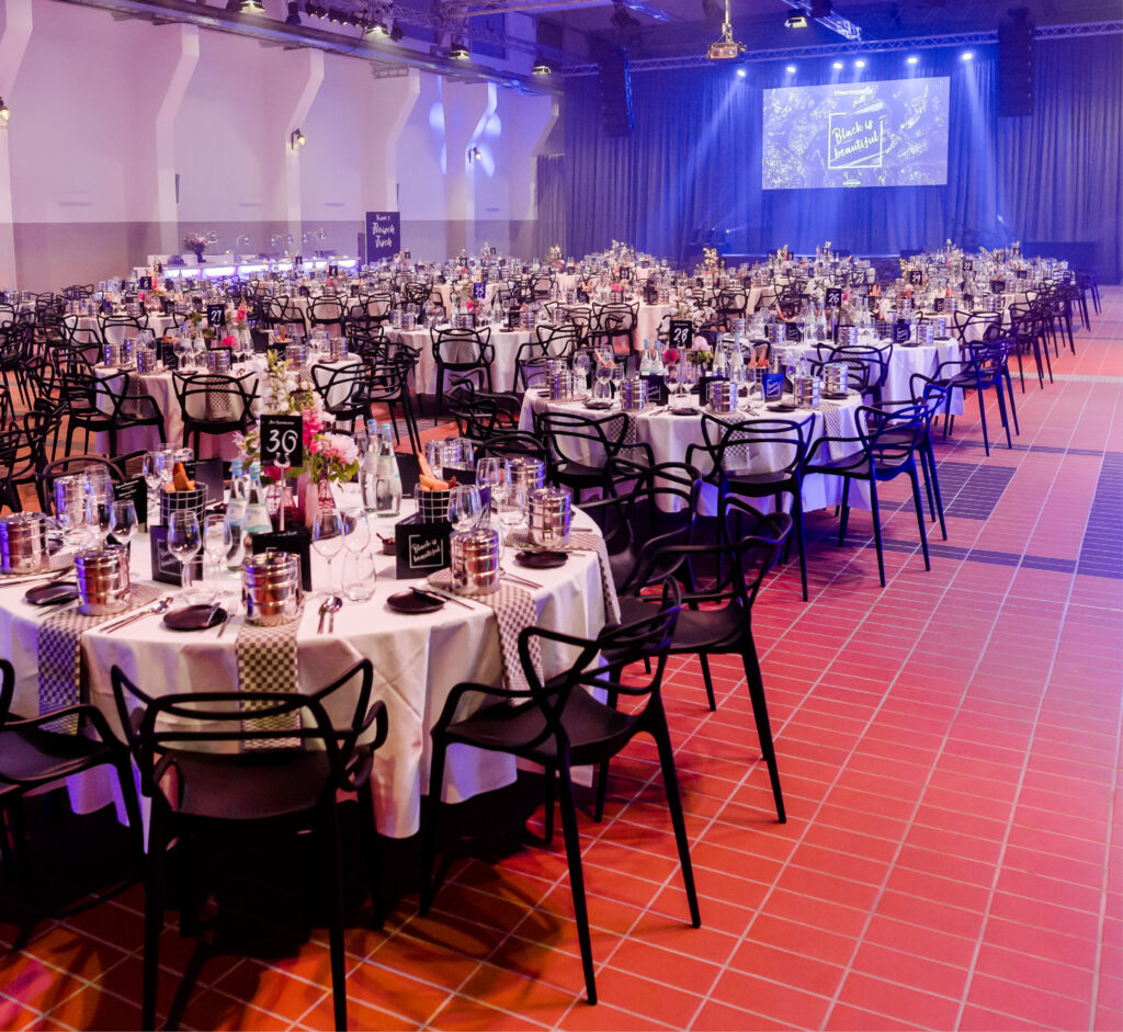 Eleganter Bankettsaal mit runden, eventgerecht gedeckten Tischen, Bühne und Leinwand im Hintergrund, violette Beleuchtung – perfekt fürs Catering in der Grand Hall Zollverein.