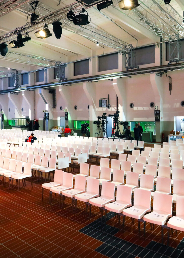 Reihen leerer weißer Stühle in einem Veranstaltungsraum mit Kameras und ein paar Leuten im Hintergrund, bereit für die Gäste, die Catering in der Grand Hall Zollverein erleben möchten.