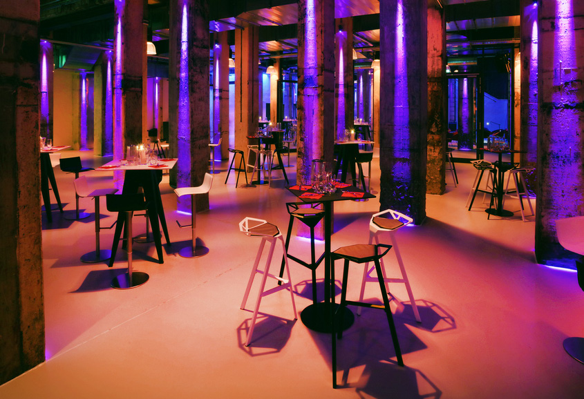 Eine moderne Loungebar mit hohen Hockern, Tischen und violetter Ambientebeleuchtung zwischen großen Betonsäulen – perfekt für Events mit Catering in der Grand Hall Zollverein.