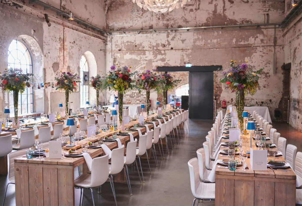 Lange, für eine Veranstaltung gedeckte Tische mit weißen Stühlen und hohen Blumengestecken in einer rustikalen Halle im Industriestil, gestaltet von Harbour Club Events und Catering.