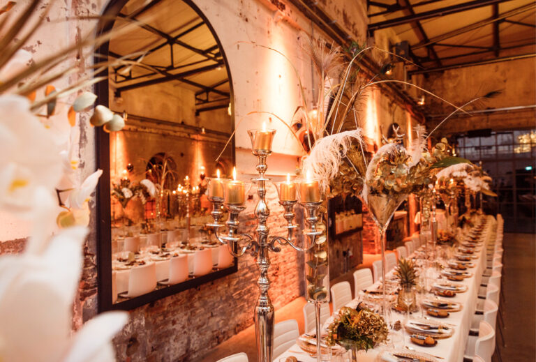 Eleganter Banketttisch mit goldenen Kerzen, Blumen und rustikalem Dekor in einem warm beleuchteten Veranstaltungsort im Industriestil von Harbour Club Events und Catering.