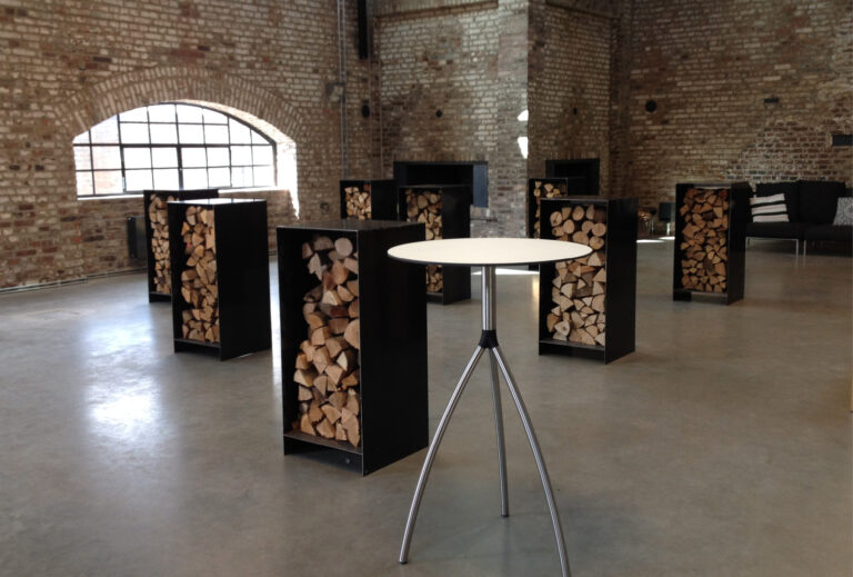 Runder Tisch mit Dreibeinfuß in einem Industrieraum, umgeben von Brennholzstapeln in schwarzen Regalen – perfekt für stilvolle Harbour Club Events und Catering-Treffen.