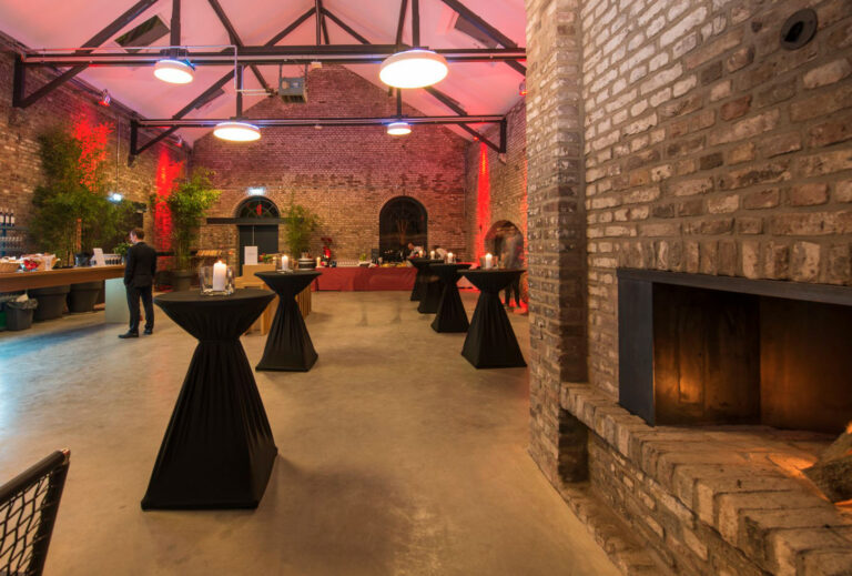 Veranstaltungsraum mit Backsteinwänden von Harbour Club Events und Catering mit Cocktailtischen, einem Buffet und einem brennenden Kamin, warm beleuchtet in Rot- und Gelbtönen.