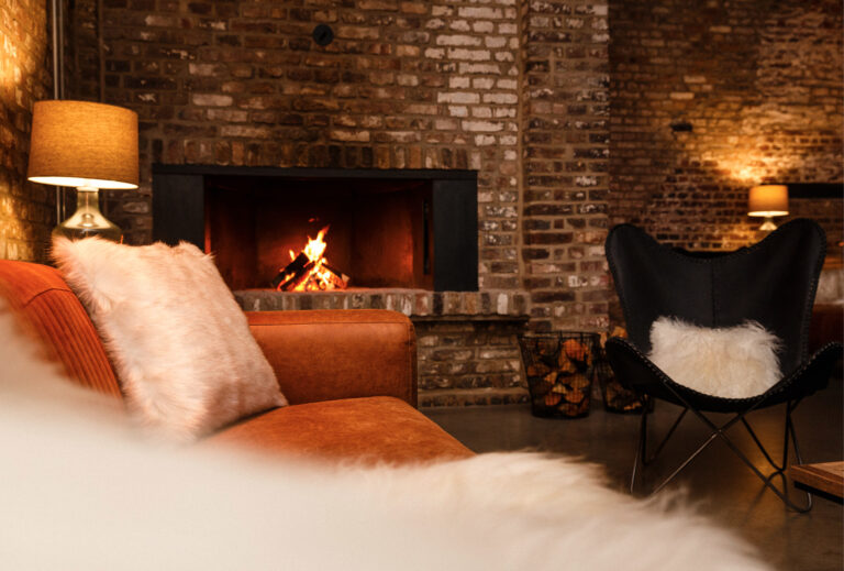 Gemütliches Wohnzimmer mit brennendem Kamin, orangefarbenem Sofa, Fellkissen und einem Schmetterlingsstuhl vor Ziegelwänden – perfekt zum Entspannen oder für die Ausrichtung von Harbour Club Events und Catering-Treffen.