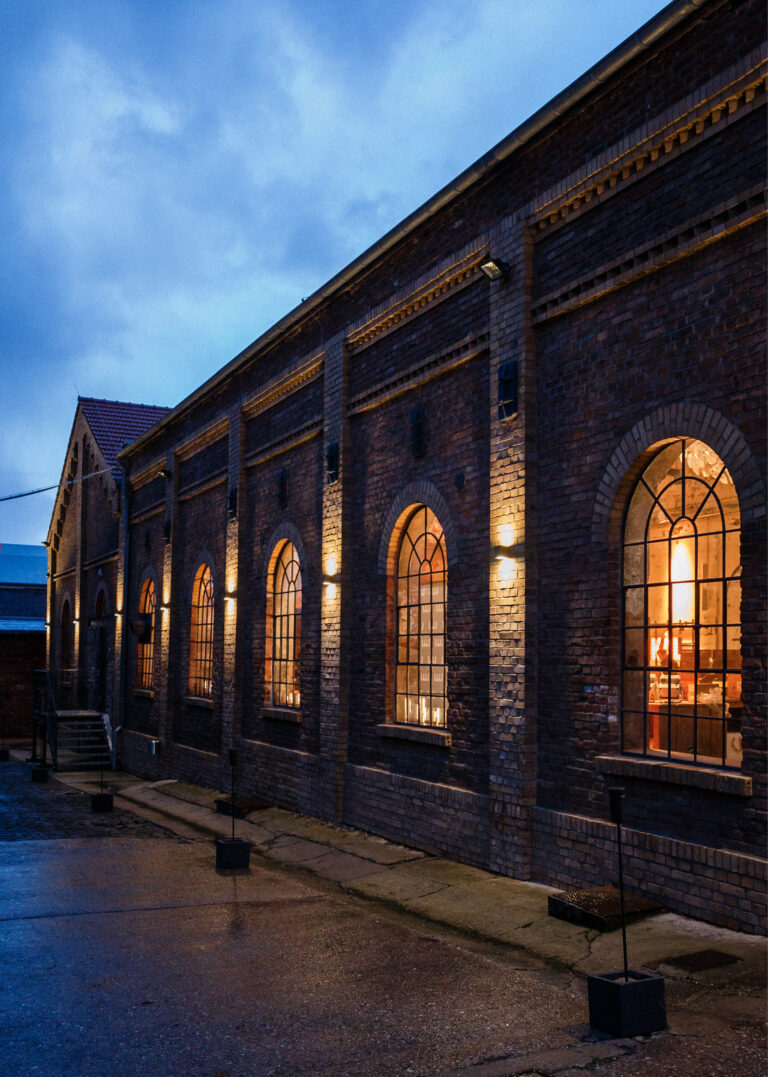 Altes Backsteingebäude mit großen Bogenfenstern, die bei Dämmerung und bewölktem Himmel von innen warm beleuchtet werden, ideal für Harbour Club Events und Catering.