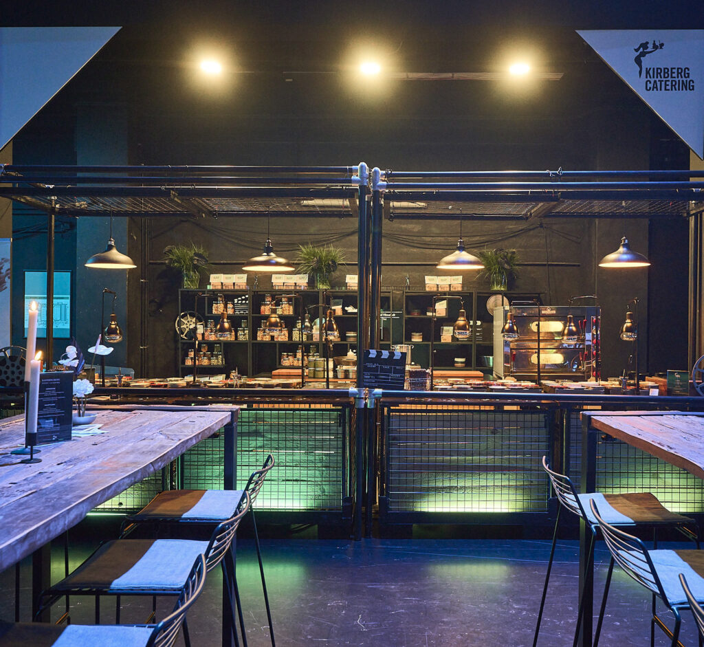 Modernes Catering-Setup mit Holztischen, Hochstühlen und einer gut beleuchteten Essenstheke im industriellen Ambiente des Studio 50 als Eventlocation.