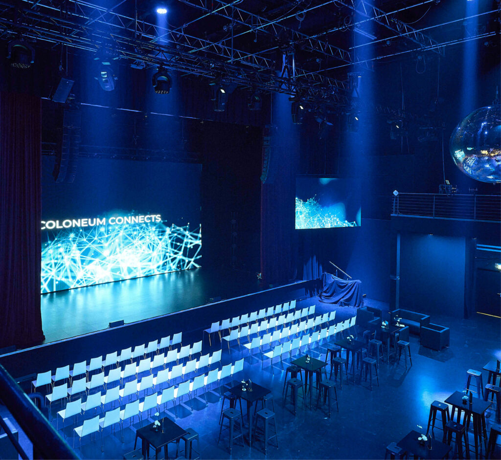 Eine Bühne mit großer Leinwand, Reihen weißer Stühle und Tische im Studio 50 als Eventlocation – einem modernen Veranstaltungsort, der durch blaues Licht in Szene gesetzt wird.