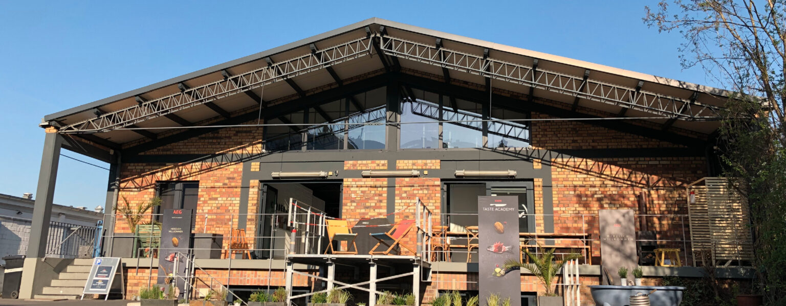 Backsteingebäude mit Metalldach, Sitzgelegenheiten auf der Terrasse und Topfpflanzen im Freien unter einem klaren blauen Himmel – perfekt für Zusammenkünfte mit Angeboten der Taste Academy Catering.