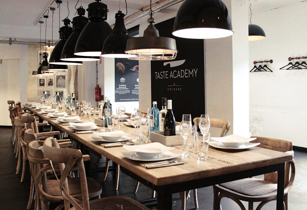 Langer Esstisch aus Holz für eine Mahlzeit in einem modernen, stilvollen Raum mit schwarzen Hängelampen – perfekt, um die Angebote der Taste Academy Catering zu genießen.