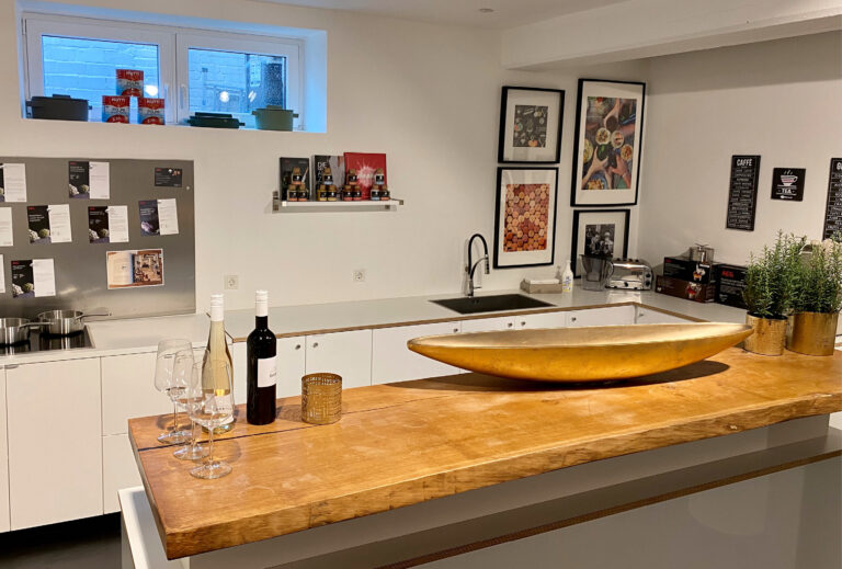 Moderne Küche mit Holzinsel, Weinflaschen, Gläsern, Kunst an den Wänden und Kräutertöpfen – perfekt zum Erkunden der Catering-Angebote der Taste Academy.