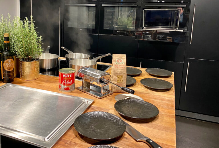 Moderne Küche mit Töpfen auf dem Herd, Nudelmaschine, Tellern, Kräutern, Tomatenkonserven und Kochutensilien auf einer Theke.