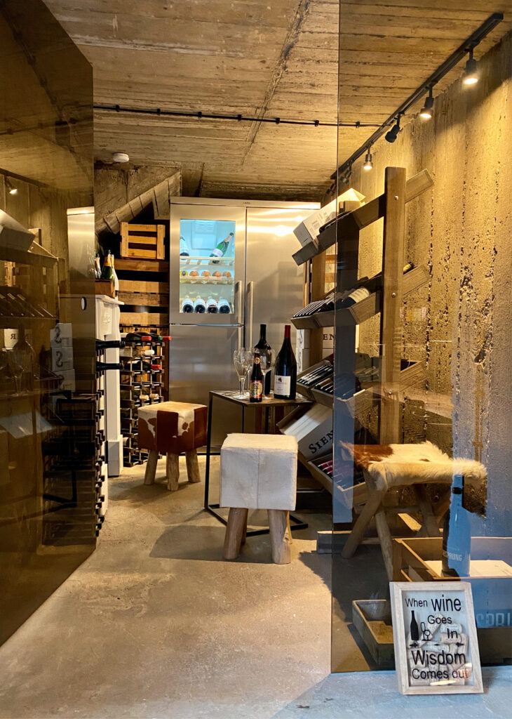 Ein moderner Weinkeller mit Holzregalen, Weinflaschen, Hockern und einem Kühlschrank im Hintergrund – perfekt für die Ausrichtung von Taste Academy Catering Angeboten.