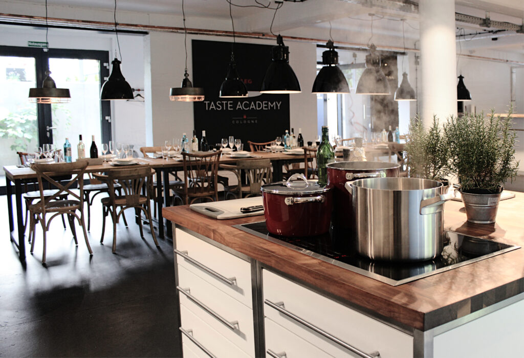Moderne Küche mit Töpfen auf dem Herd im Vordergrund, Esstische im Hintergrund unter Pendelleuchten, Präsentation von Taste Academy Catering Angeboten für jeden Anlass.