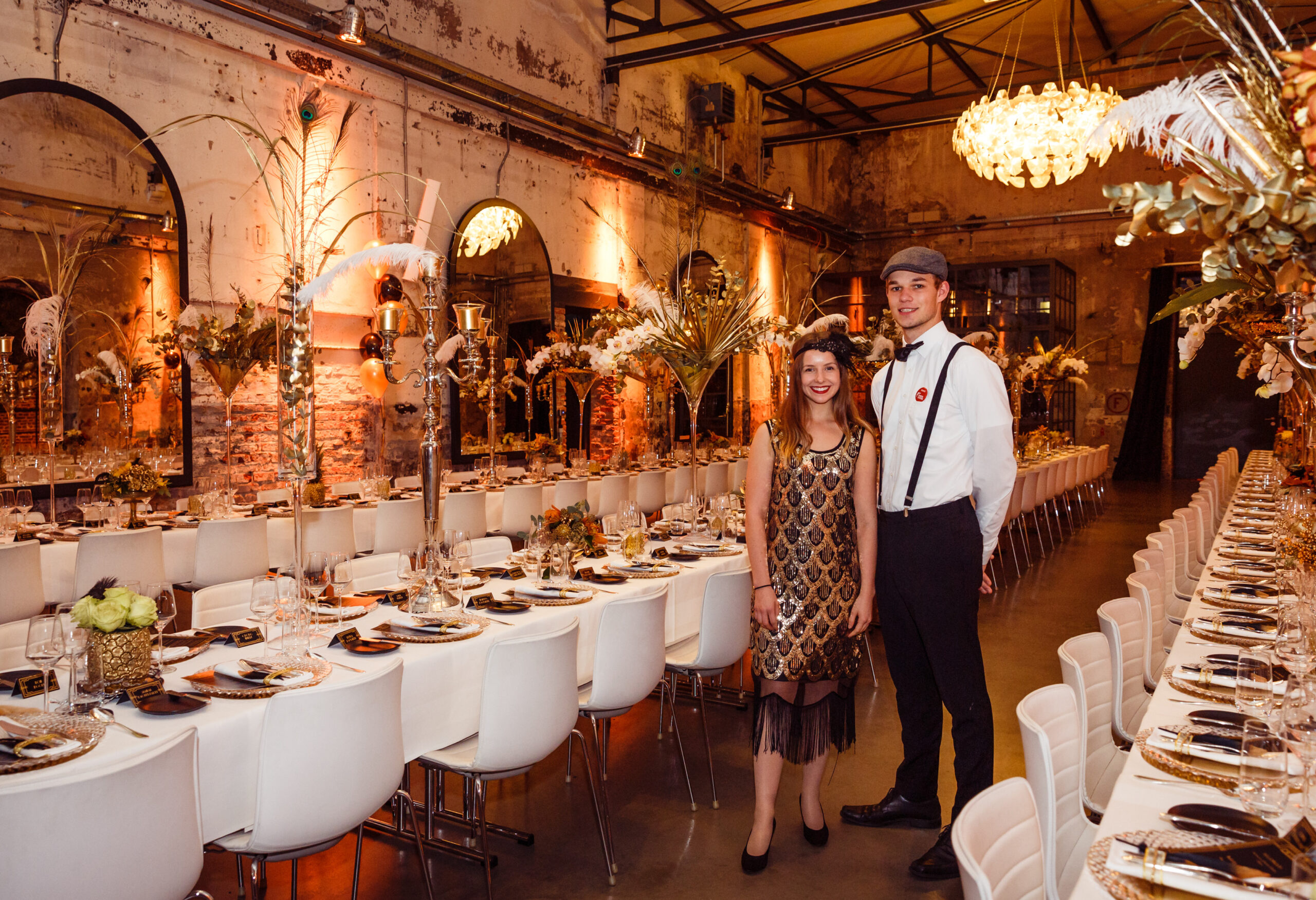 Zwei Personen in 1920er-Jahre-Outfits stehen in einem aufwendig dekorierten, langen Bankettsaal mit elegant gedeckten Tischen – perfekt für Event Catering für besondere Anlässe.