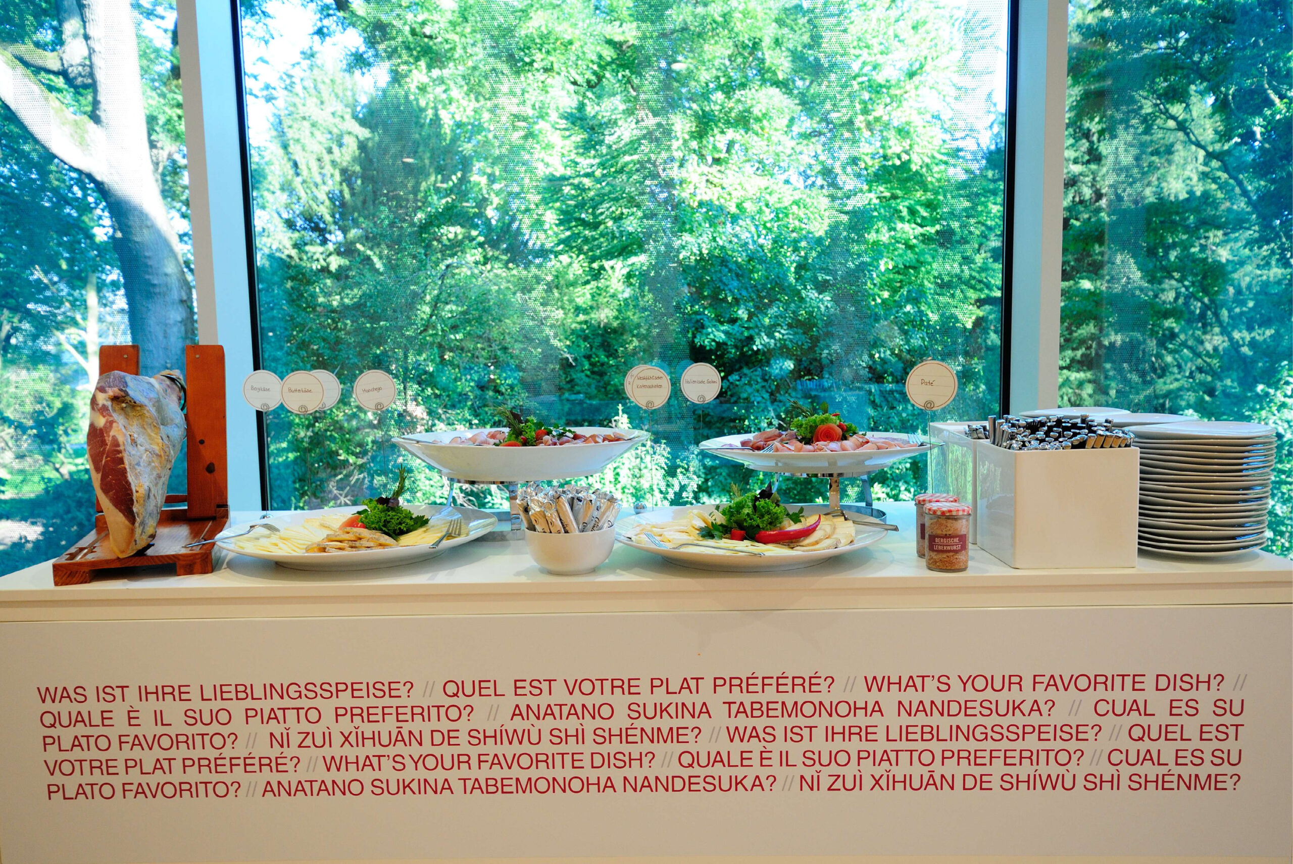 Buffettisch mit Salaten, Tellern und Besteck in der Nähe eines Fensters mit der Aufschrift „Was ist Ihr Lieblingsgericht?“ in mehreren Sprachen – perfekt für das Catering für Tagungen und Konferenzen.