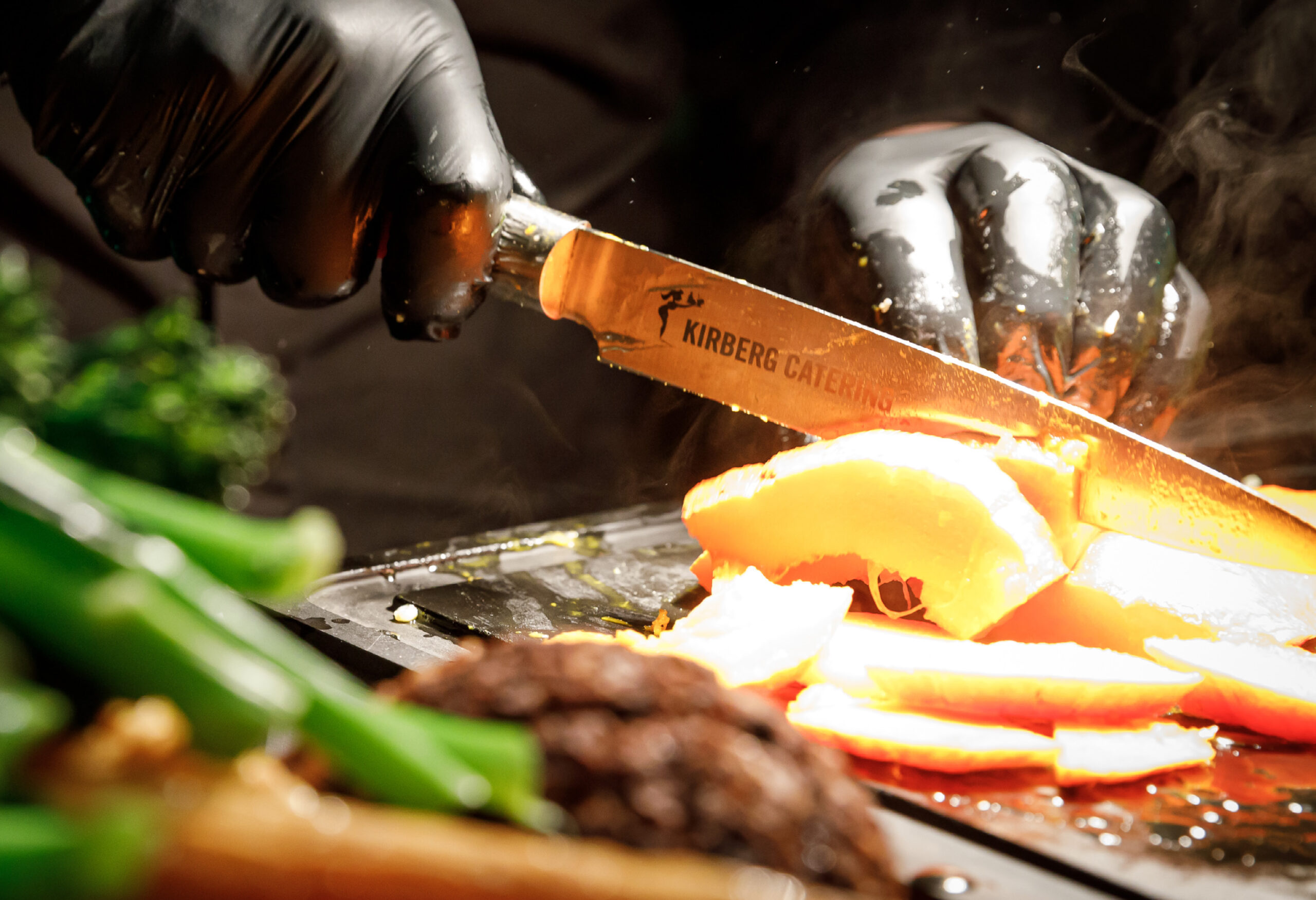 Ein Koch in schwarzen Handschuhen schneidet Gemüse mit einem Messer auf einem dampfend heißen Grill.