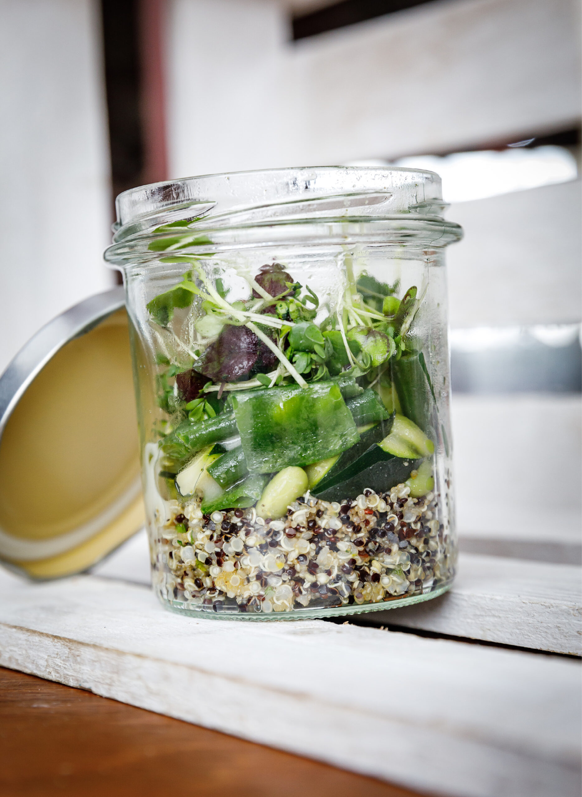 Ein Glasgefäß gefüllt mit Quinoa, Gurke, Gemüse und Sprossen steht auf einer weißen Holzoberfläche.